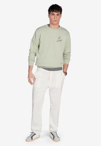 Sudadera verde claro con cuello redondo y detalle de logo, combinada con pantalones blancos de corte relajado y zapatillas casuales con acentos azules.