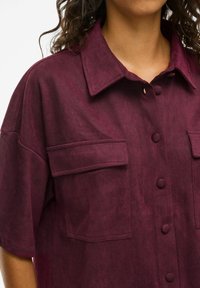 Bordo krāsas pogas aizdares krekls ar teksturētu audumu, ar izplatītu apkakli, divām krūšu kabatām un lielām pogām. Īsas piedurknes un brīvs piegriezums.