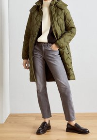Manteau matelassé vert olive avec capuche, superposé sur un pull crème et foncé, associé à un jean gris et des mocassins noirs. Tissu texturé.