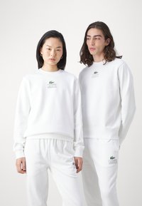 Fehér pamut kapucnis pulóverek és melegítőnadrágok zöld Lacoste logóval. A laza szabás bordás mandzsettákkal és kerek nyakkivágással rendelkezik.