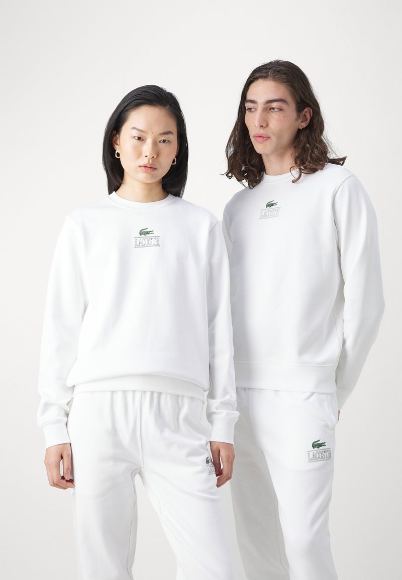 Felpe e pantaloni tuta in cotone bianco presentano un logo Lacoste verde. La vestibilità comoda include polsini a costine e un colletto a girocollo.