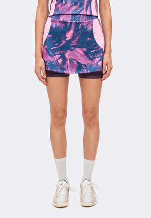 Femme portant une jupe de sport rose et marine avec des shorts intégrés, associée à des chaussettes blanches et des baskets blanches, debout les mains détendues le long du corps.