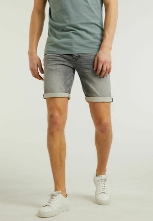 Homme portant un short en jean gris retroussé, un t-shirt vert et des baskets blanches, debout devant un fond uni.