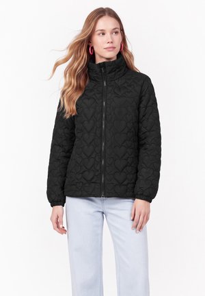Femme portant une veste matelassée noire avec des motifs en forme de cœur, un jean bleu clair et des boucles d’oreilles roses en forme d’anneau, debout devant un fond blanc.