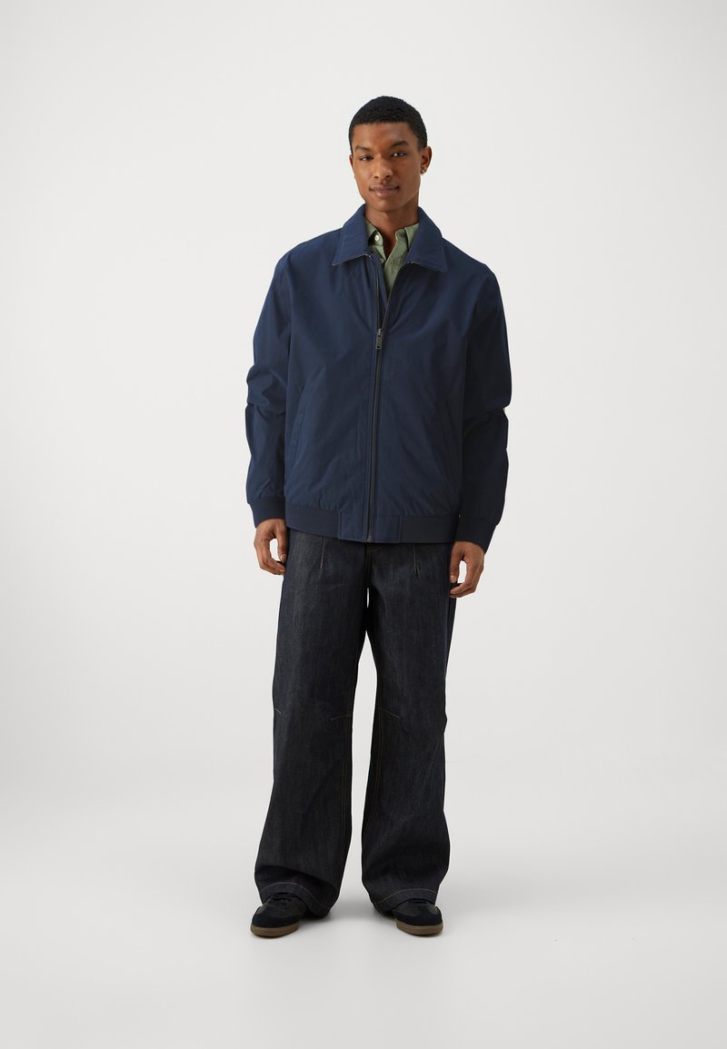Giacca bomber blu navy con zip frontale, vestibilità leggermente oversize e colletto a punta, indossata sopra una camicia verde e jeans scuri.