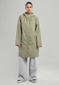 Vihreä vedenpitävä takki hupulla, napituksilla, sivutaskuilla ja suoralla leikkauksella, yhdistettynä harmaisiin leveisiin housuihin ja mustiin lenkkareihin.