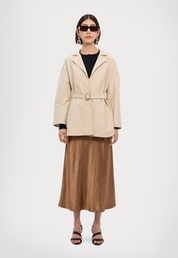 Beige oversized blazer med belte, kombinert med en bronsefarget satin midi-skirt. Figuren bærer svarte solbriller og store hoop-øreringer.