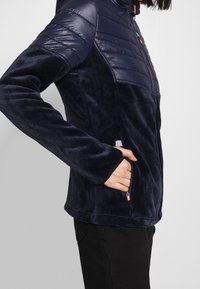 Nainen, joka on pukeutunut tummansiniseen vetoketjulliseen takkiin, jossa on palotetut hartiat ja pehmeä kangasosa, kädet vetoketjullisissa sivutaskuissa, yksinkertaista taustaa vasten.