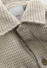 Beige textured fabric with a grid pattern, featuring a fold-down collar and a brown button. Label reads "NEXT."