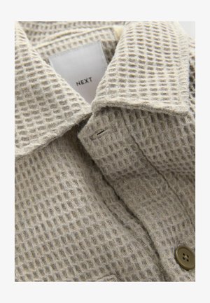 Bežs teksturēts audums ar režģa rakstu, ar nolokāmu apkakli un brūnu pogu. Uz birkas ir rakstīts "NEXT".