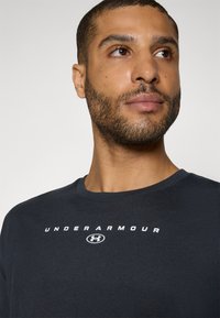Unter Armour schwarzes T-Shirt mit rundem Halsauschnitt, versehen mit weißem Logo und Schriftzug über der Brust. Weiche, glatte Stoffstruktur.