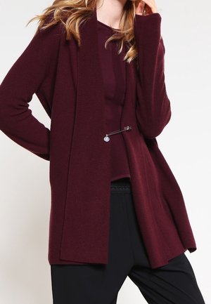 Osoba nosící tmavě vínový cardigan přes sladěný top, upevněný ozdobnou sponou, v kombinaci s volnými černými kalhotami.