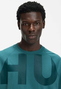 Morska bluza z dużym, ciemnozielonym napisem „HU”, okrągłym dekoltem i rękawami raglanowymi o gładkiej fakturze.