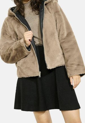 Vrouw draagt een beige nepbontjas over een bruine trui met een zwarte rok, en houdt de jas met één hand open.