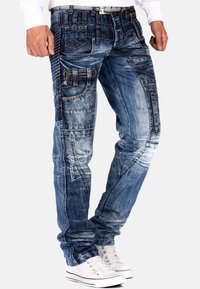 Jeans in denim blu da uomo con finitura usurata, multiple tasche e cuciture decorative; abbinati a sneakers bianche.