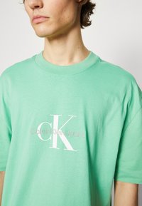 Camiseta de algodón verde menta con cuello redondo, que presenta un logotipo blanco "CK" y el texto "Calvin Klein Jeans" impreso en la parte frontal. Mangas cortas.