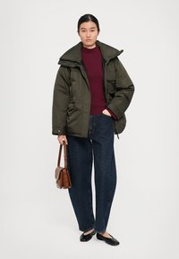 Woman standing wearing olive green puffer jacket, burgundy sweater, dark blue jeans, black flats, holding brown handbag by her side.