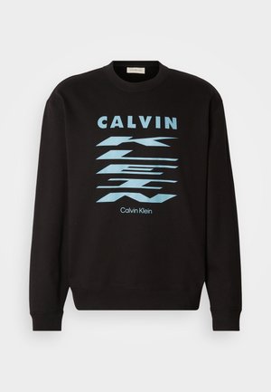 Črna majica s kapuco s krožnim izrezom, ki vsebuje modro grafično obliko in napis "CALVIN KLEIN." Rjavkasti manšeti in rob. Mehka tekstura.