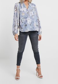Blusa floreale azzurro chiaro con maniche a palloncino, abbinata a jeans skinny scuri e sandali con tacco bianco. Dettaglio delicato sullo scollo.