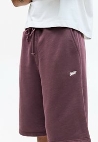Shorts en coton bordeaux avec cordon de serrage, poches latérales, bords côtelés et un logo blanc sur la jambe gauche. Texture douce et coupe décontractée.