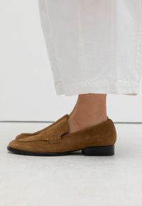 Mocassins en suede marron avec un petit talon noir, présentant un motif cousu et un rabat à l'avant. Associés à un pantalon blanc légèrement cropped.