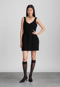 Robe noire sans manches avec un décolleté en V, un buste structuré et une jupe évasée. Portée avec des bas noirs transparents montants et des escarpins à bout pointu.
