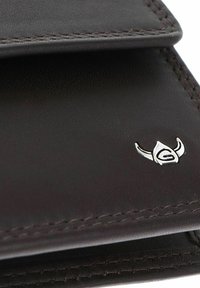 Gros plan sur un coin de portefeuille en cuir marron foncé avec des coutures détaillées et un petit logo casque viking argenté sur la surface.