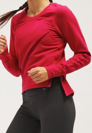 Kvinde iført en skrigrød langærmet træningstrøje og sorte leggings, poserer som om hun jogger eller løber.