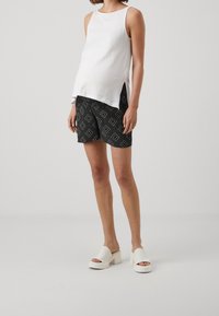 Bílý bezrukávový top s bočními rozparky, vzorované černé kraťasy s geometrickými vzory a mohutné bílé sandály na klínu.