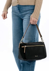 Black handbag made of smooth material, featuring a zippered closure, two zip pockets, and brown accents. Worn by a person with blue jeans.