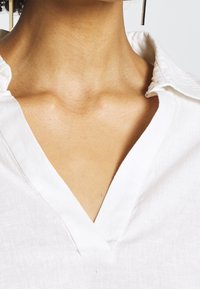 Blusa bianca in lino con scollatura a V, caratterizzata da un tessuto testurizzato e un colletto con dettagli di cucitura discreti.