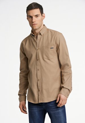 Camisa de botones marrón claro con un bolsillo en el pecho, textura sutil y mangas largas. Combinada con jeans azul oscuro.