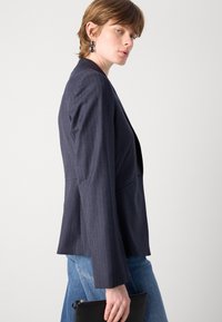 Námořnicky pruhovaný blazer s přiléhavým střihem, klopovými límci a zapínáním na jeden knoflík. S kombinací s světle modrými džíny a držením černé kabelky.