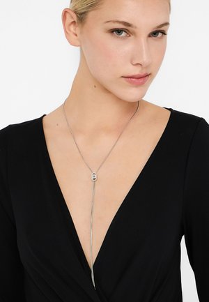 Silberne Halskette mit langer Kette, rundem, verziertem Anhänger und zwei verlängerten Strängen, getragen über einem schwarzen Kleidungsstück.