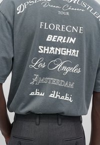 T-shirt grigio scuro con stampa bianca del tour che elenca città come Firenze, Berlino, Shanghai, Los Angeles, Amsterdam e Abu Dhabi.