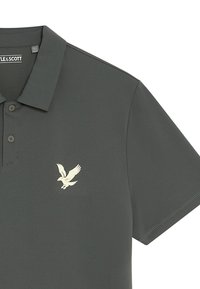 Polo shirt grigio scuro realizzato in tessuto morbido, con colletto corto e tre bottoni, e logo di un'aquila bianca sul lato sinistro del petto.