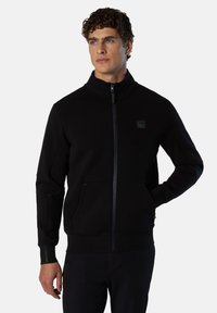 Felpa nera con zip, realizzata in tessuto morbido, presenta un colletto alto, tasche laterali e una patch con logo sul petto. Include fori per il pollice sui polsini.