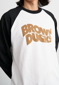 Weißes Sweatshirt mit schwarzen Raglanärmeln, versehen mit großem braunem Text "BROWN DUCKS", der ein mutiges, grafisches Design zeigt.