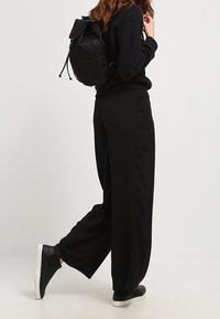 Osoba oblečená v černých širokých kalhotách, černé mikině, černých teniskách s bílými podrážkami a černém batohu, stojí na jedné noze.