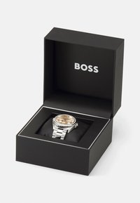 Montre en argent avec un cadran doré, présentée dans une boîte noire avec un intérieur doux. Comprend un bracelet en métal et une lunette détaillée. Nom de la marque "BOSS" visible.