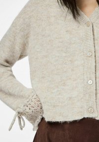 Beige buttoned cardigan with textured knitting and lace-up cuffs worn over a brown skirt, showing upper torso and arm.