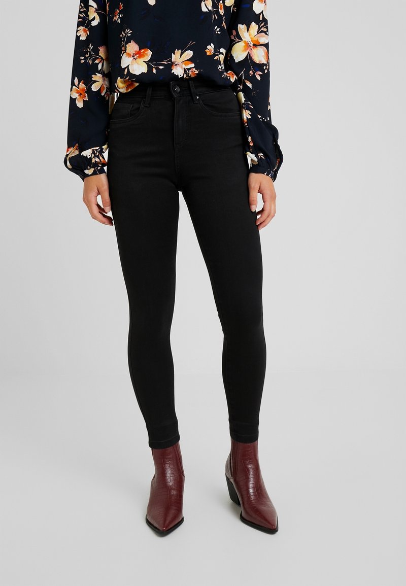 Femme portant un jean skinny noir, un chemisier floral foncé et des bottines bordeaux à bout pointu, debout devant un fond uni.