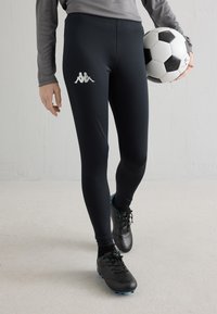 Oseba v črnih športnih pajkicah in kopačkah drži črno-belo nogometno žogo na enostavni svetli podlagi.