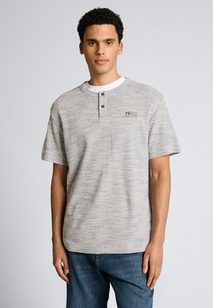 Młody mężczyzna z krótkimi kręconymi włosami, ubrany w jasnoszary, krótki rękaw henley oraz niebieskie dżinsy, stojący na jednolitym tle.