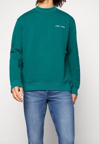 Osoba v modrozelené mikině s malým bílým logem a světle modrých džínách stojí před jednoduchým světlým pozadím.