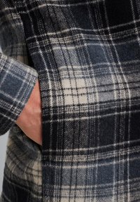 Ruudullinen villakangastakki, jossa on tummansinisen ja kermanvärin sekoitus, pehmeä rakenne ja sivutasku, jossa on käsi sisällä.