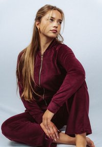 Burgundowy welurowy dres na zamek z długimi rękawami, spodnie o luźnym kroju i gładka tekstura. Modelka siedząca, dłonie spoczywają na kolanach.