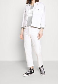 White denim jacket over a gray sweatshirt with “TOMMY HILFIGER” text, paired with white cropped pants and black high-top sneakers.