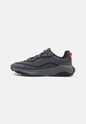 Ciemnoszary sneaker z fakturowaną podeszwą, sznurowadłami, czerwonymi oczkami, wyściełanym kołnierzem i czerwoną pętlą na pięcie, pokazany na białym tle.