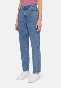 Mulher a usar calças de ganga azul de perna reta de cintura alta e ténis brancos, a apoiar-se contra um fundo liso.